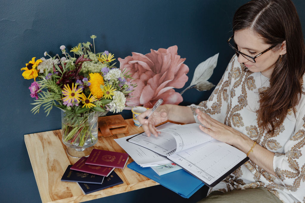 Anna Aresi at her working desk. On the desk are a planner, some papers and two Italian and two US passports and a vase of colorful cut flowers.