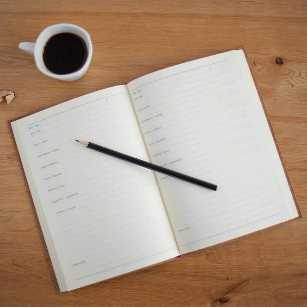 A multilingual Italian-English planner is spread open on a table. There is a pencil across the page and a cup of coffee next to it.