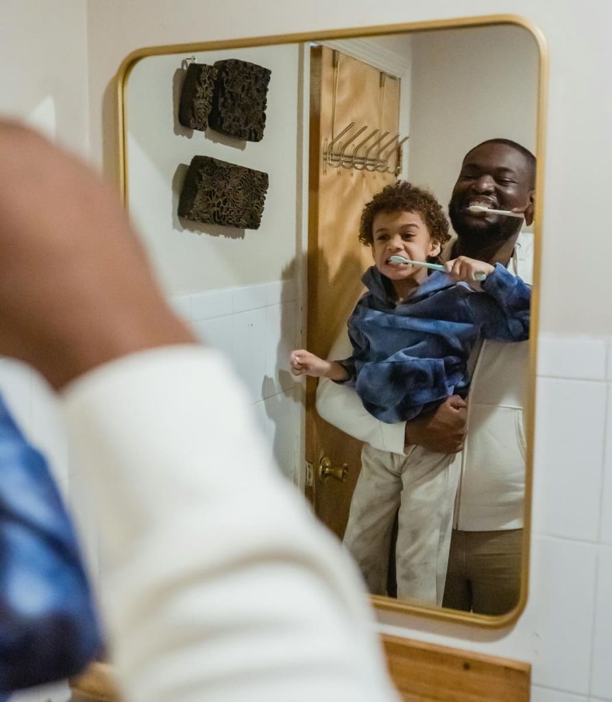 A black dad and his son brush their teeth together in front of the mirror