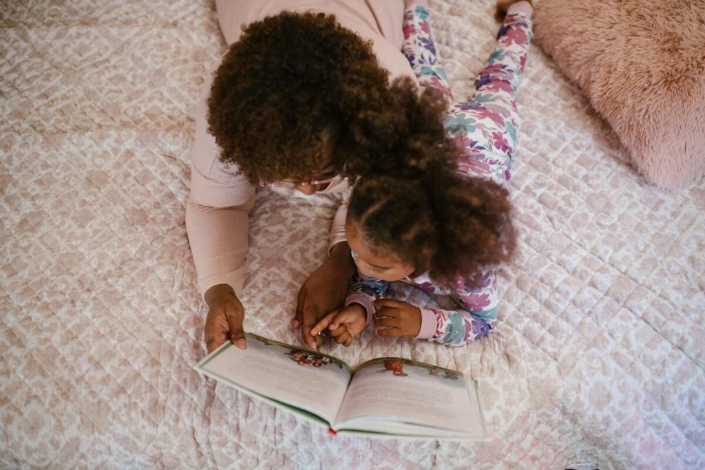 A black mom and her daughter read a book together. The daughter is pointing to a word or image and saying something or trying to read.