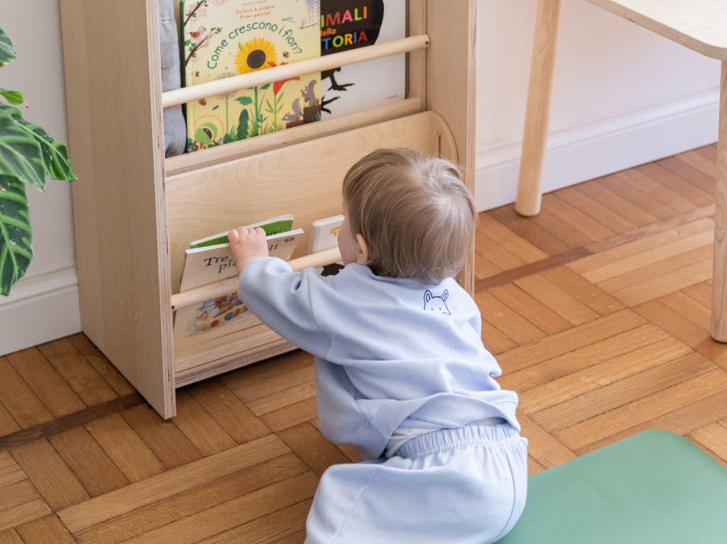 A white toddler pulls out a book from a Montessori bookcase. 